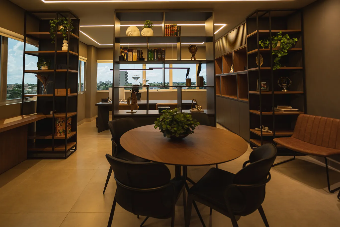 Interior sofisticado de escritório de advocacia com estantes amplas em estrutura metálica preta e prateleiras de madeira repletas de livros jurídicos, objetos decorativos, troféus e plantas, mesa redonda de madeira ao centro com cadeiras estofadas escuras e poltrona de couro caramelo ao fundo, iluminação quente em fitas de LED no teto formando linhas geométricas, janelas amplas com vista da cidade ao fundo, paleta em tons de marrom, bege e dourado, atmosfera profissional e acolhedora, fotografia arquitetônica ultra-realista